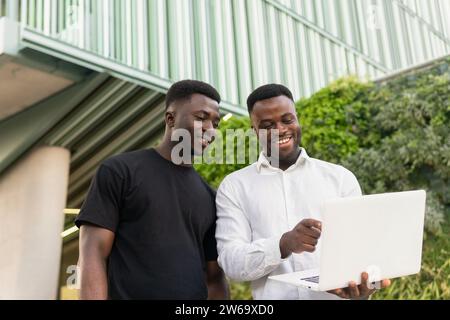 Low angle of cheerful African American businessman pointing at netbook screen while showing project to male colleague outside company building Stock Photo