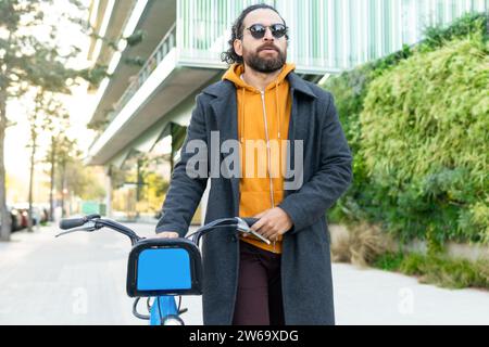 Stylish man in a coat and yellow hoodie walking with his bike in an urban setting. Stock Photo