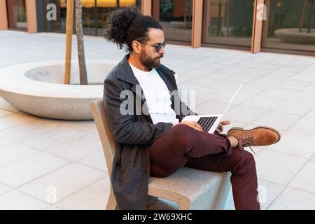 Man with a laptop working outdoors on a modern urban bench. Stock Photo