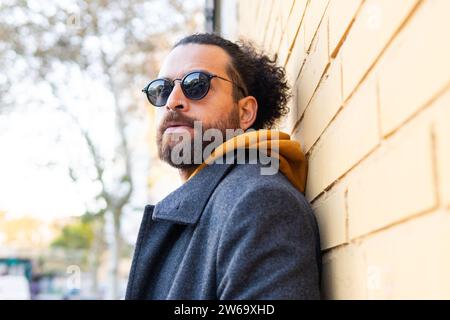 A trendy man sporting sunglasses and a casual, yet fashionable outfit with a scarf, leans confidently against a brick wall outside. Stock Photo