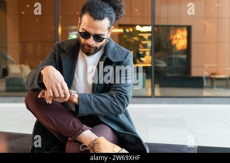 A fashionable man in casual business attire is sitting outside an urban building, looking at his watch. Stock Photo