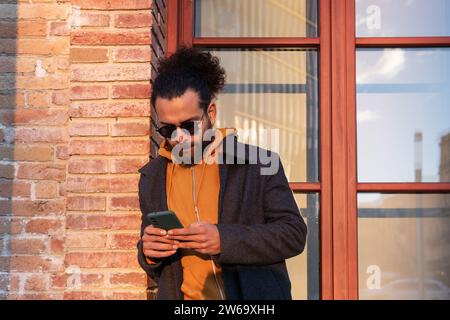 A trendy young man in sunglasses and a warm coat is focused on texting on his smartphone, standing by a red brick wall with sunshine lighting his side Stock Photo