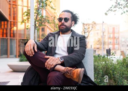A fashionable man with a beard and sunglasses sits casually in a cityscape, exuding confidence and style. Stock Photo