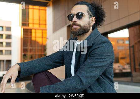 A trendily dressed man with sunglasses and a beard sits casually in an urban environment during the daytime, exuding confidence and style. Stock Photo