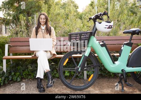 A young woman sits on a bench in a sunny park, typing on her laptop next to her parked electric bicycle. Stock Photo