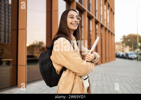 A cheerful young woman with a backpack standing outside a modern building, holding a notebook and looking optimistic. Stock Photo