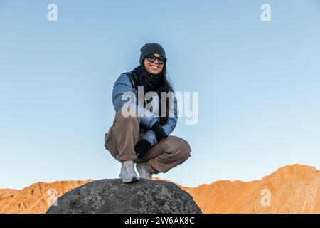 From below of female tourist with toothy smile in sunglasses and warm clothes looking at camera while kneeling down on rock against shining yellow mou Stock Photo