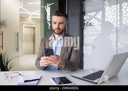 Serious concentrated mature businessman thinking reading online social networks, experienced mature man holding phone using smartphone at workplace inside office with laptop. Stock Photo