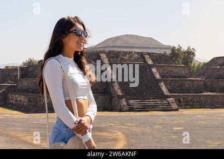 Side view of smiling brunette tourist in casual clothes with sunglasses and handbag standing at historic construction in Mexico Stock Photo