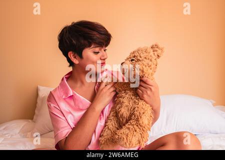 Side view of smiling young woman sitting on comfortable bed and holding small teddy bear in bedroom Stock Photo