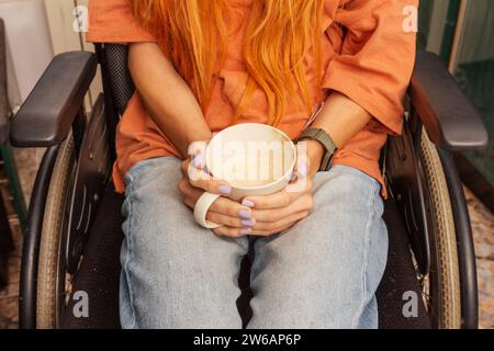 Crop os a person sits in a wheelchair, cradling a white coffee cup in their hands, wearing casual clothes. Stock Photo