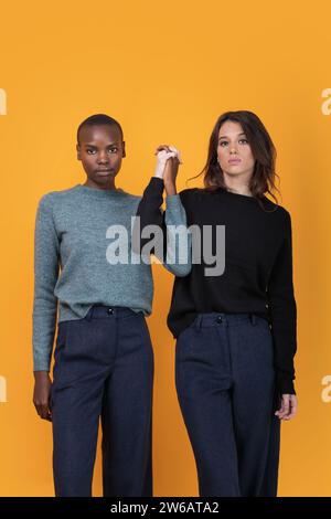 Confident multiracial young female friends in trendy casuals holding hands while standing looking at camera against yellow background Stock Photo