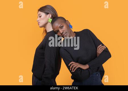 Side view of diverse young female friends leaning on each other looking at camera while wearing trendy colorful earrings against yellow background Stock Photo