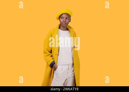 Thoughtful fashionable African American young woman wearing flat cap and long coat while looking away against yellow background Stock Photo