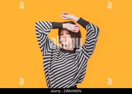 Beautiful calm young slim Caucasian woman in trendy casuals looking down while hands on head against yellow background Stock Photo