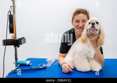 Smiling young female groomer in black clothes embracing cute American cocker spaniel while leaning on blue table against white background in grooming Stock Photo