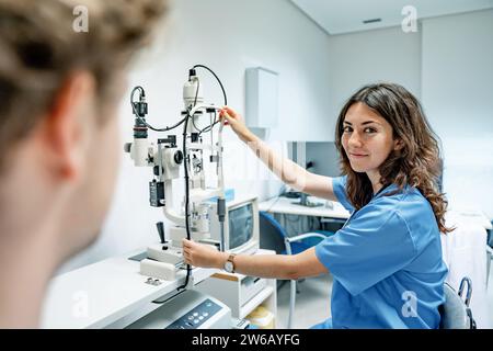 Smiling young female doctor in uniform and stethoscope looking at camera while standing with hand on slit lamp biomicroscope in hospital under illumin Stock Photo
