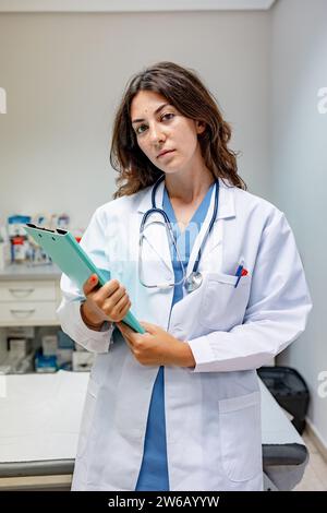 Young female doctor wearing uniform and stethoscope standing with folder in hospital while looking at camera against white wall Stock Photo