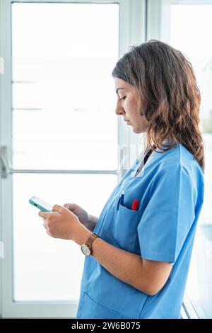 Side view of young female doctor wearing blue uniform standing in modern hospital and browsing mobile phone Stock Photo