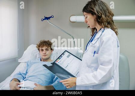 Side view of young female doctor with clipboard and stethoscope standing near patient lying on bed in hospital and checking documents while looking at Stock Photo