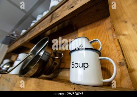 Low angle of white soup mugs stacked on a rustic wooden kitchen shelf, with metal pots hanging below, creating a cozy and organized culinary setting Stock Photo