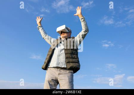 From below joyful elderly man with arms raised wearing a VR headset in an open field Stock Photo