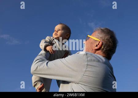 From below cheerful elderly man with sunglasses lifting a baby up in the air, both looking away with a clear blue sky above Stock Photo