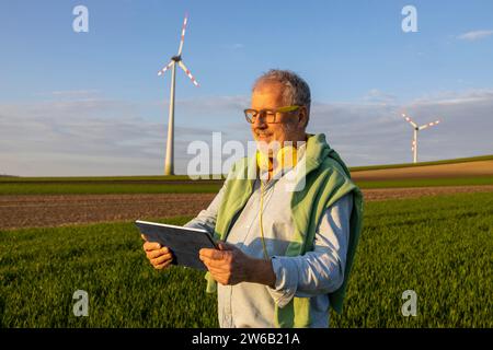 Happy senior man with headphones around neck using digital tablet at wind farm against blue sky Stock Photo