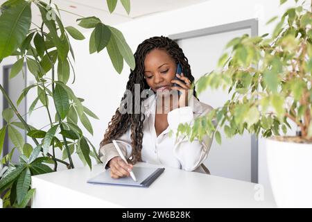 Young African American businesswoman using smartphone while writing with stylus on digital tablet at desk in office Stock Photo