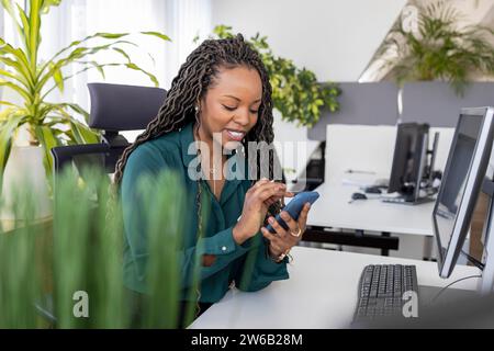 Happy young African American businesswoman using smartphone while sitting at desk in office Stock Photo