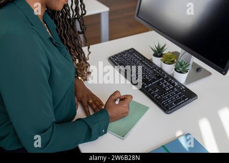 From above of young African American businesswoman writing on notepad at computer desk in office Stock Photo