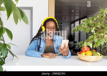 Happy young African American businesswoman using headphones and smartphone at desk in creative office Stock Photo