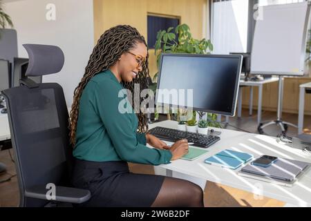 Side view of young happy African American businesswoman writing on notepad at computer desk in office Stock Photo