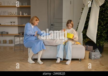 A cozy scene of a mother and daughter sitting on a footstool, wrapping Christmas gifts by a decorated tree. Stock Photo
