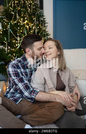 Young couple in a loving embrace, with the man affectionately holding the woman as they sit in front of a twinkling Christmas tree at home Stock Photo