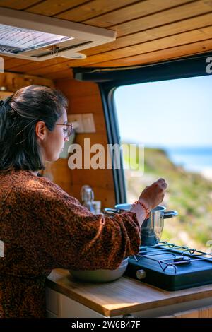Side view of focused female traveler in casual attire and eyeglasses cooking food on stove by window in modern kitchen during trip in camper van Stock Photo
