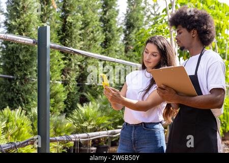 Young Hispanic agronomist showing smartphone to African American coworker standing with clipboard while working together in greenhouse Stock Photo
