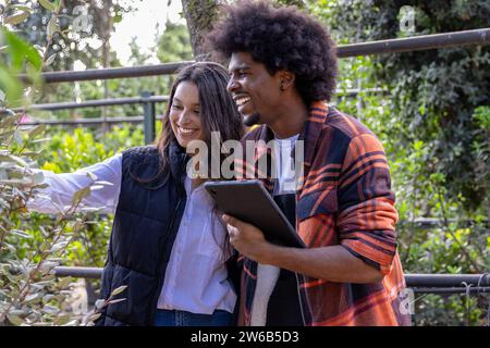 Smiling young diverse farm coworkers dressed in jackets using digital tablet while standing at organic greenhouse Stock Photo