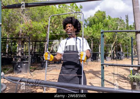 Happy young African American male farmer with closed eyes carrying metal rack while working at organic greenhouse against trees on cloudy day Stock Photo