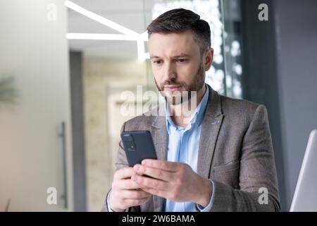Serious concentrated mature businessman thinking reading online social networks, experienced mature man holding phone using smartphone at workplace inside office with laptop. Stock Photo