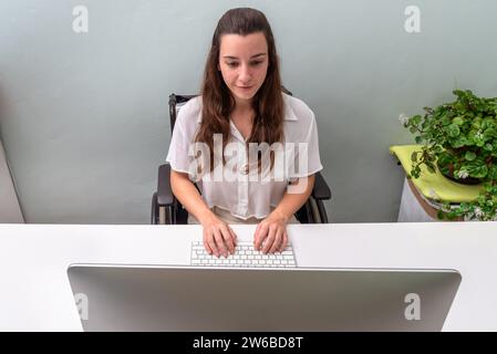 A focused young professional woman types on a keyboard at a tidy workspace with a potted plant nearby. Stock Photo