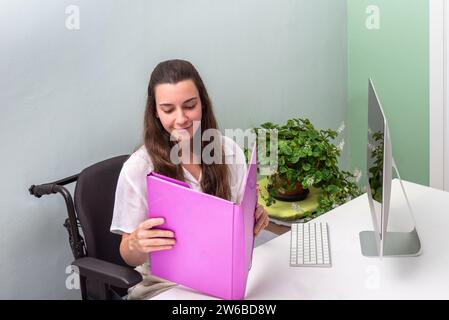 A young woman in wheelchair in a casual white blouse is focused on reading a bright pink folder while at her workspace with an adjacent potted plant a Stock Photo