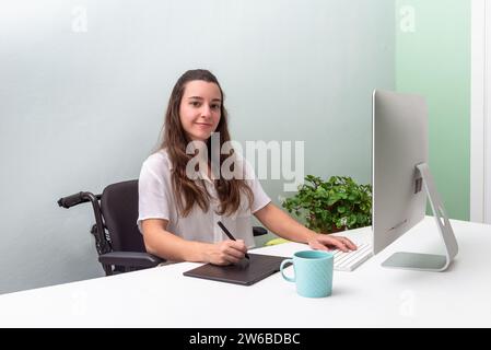 A focused professional woman sits in wheelchair at a clean, minimalist desk, working on a computer with a stylus on a digital tablet, in a well-lit of Stock Photo