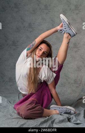 A young woman in casual attire performs a flexible yoga pose, stretching her leg upwards while seated on a draped fabric Stock Photo