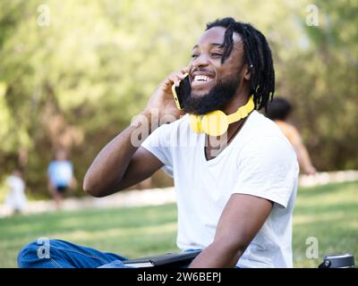 Cheerful young African American guy with headphones around neck sitting on lawn in sunny park and talking via mobile phone Stock Photo