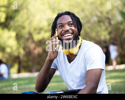 Cheerful young African American guy with headphones around neck sitting on lawn in sunny park and talking via mobile phone Stock Photo