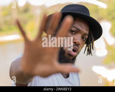 Young African American bearded man with dreadlocks in black hat outstretching hand towards camera and showing stop gesture Stock Photo