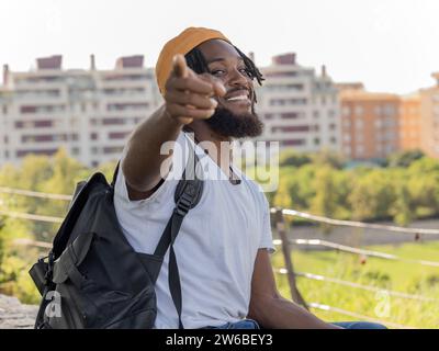 Smiling young African American hipster in yellow hat with backpack sitting against city buildings and pointing at you Stock Photo