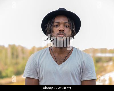 Young African American bearded guy with dreadlocks in hat and white t shirt looking at camera with serious glance against blurred nature Stock Photo