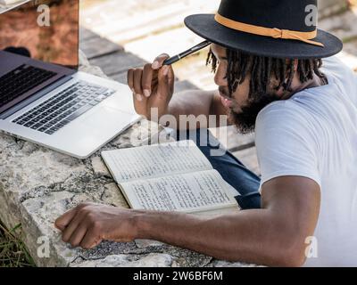 High angle of young African American male blogger in hat sitting on ground touching forehead with pen while reading notes and thinking in park with la Stock Photo